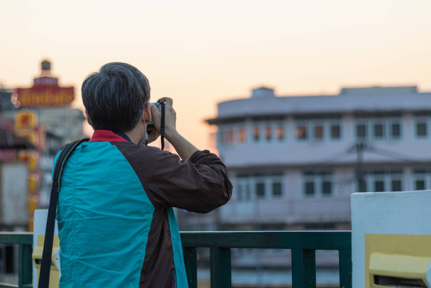 Un anciano usando una cámara réflex digital para capturar imágenes de una ciudad turística de Tailandia solo en la noche antes de que se ponga el sol.El concepto de una persona que tiene que vivir sola cuando es mayor - Foto, Imagen