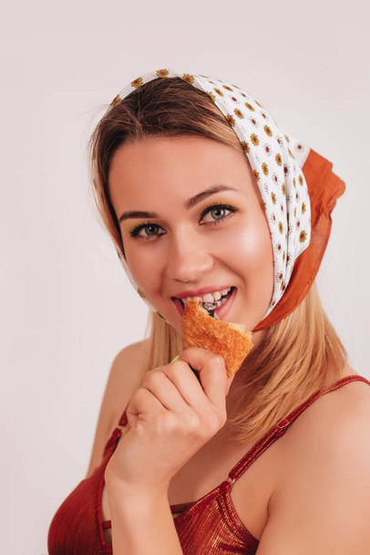 Élégante fille souriante dans un body brun, un jean et une écharpe sur sa tête en mangeant un croissant frais sur un fond blanc. La fille prend une bouchée du croissant.  - Photo, image