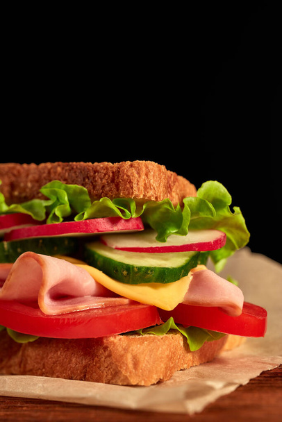 Appetizing sandwich from ham, cheese and vegetables serving on parchment paper is laying on wooden table on black background with copy space. - Foto, afbeelding