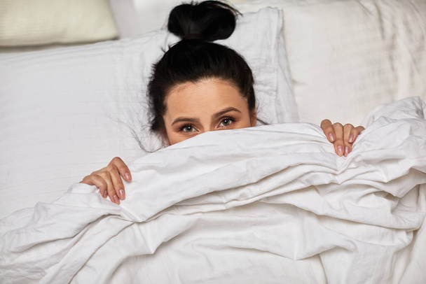 Playful cute young woman is lying in bed and hiding under blanket in the morning - Foto, Bild