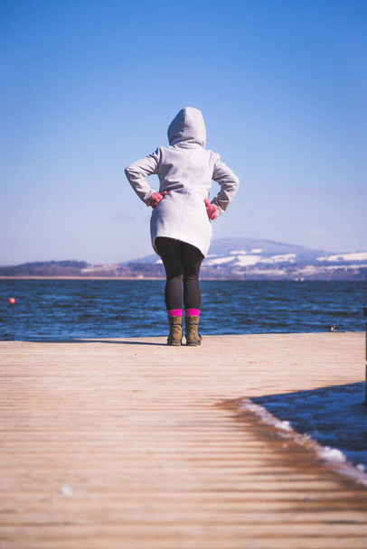 Young girl with grey coat is standing on a footbridge and enjoys the view over the lake, winter time - Fotó, kép