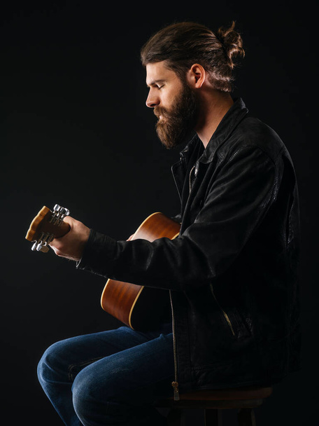 Young bearded man sitting and playing an acoustic guitar over black background. - Foto, afbeelding