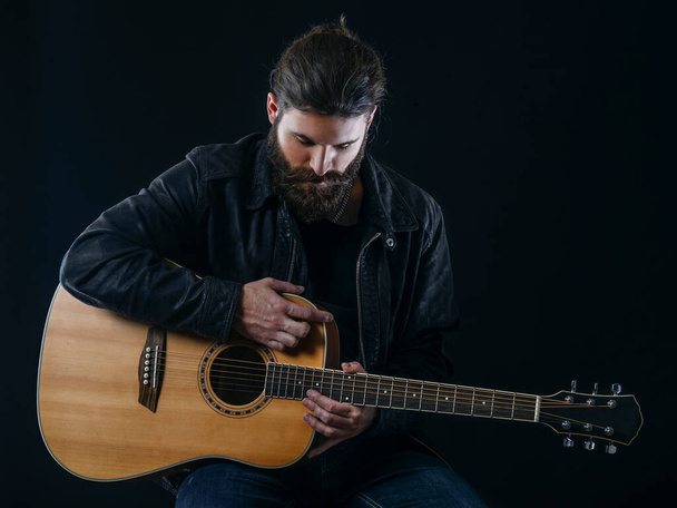 Young bearded man sitting and playing an acoustic guitar over black background. - Foto, imagen