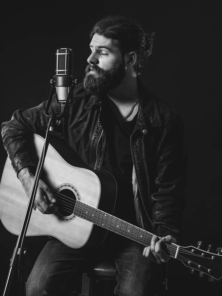 Young bearded man sitting, playing an acoustic guitar and singing over black background. - Valokuva, kuva