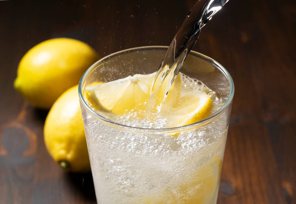 Pour soda water into a glass of lemon placed against a dark wooden background. Image of a lemon cocktail - Φωτογραφία, εικόνα