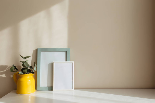 A yellow ceramic jug or vase with eucalyptus branches, empty white photo frames on the white table in the interior with beige walls near window. - Фото, изображение