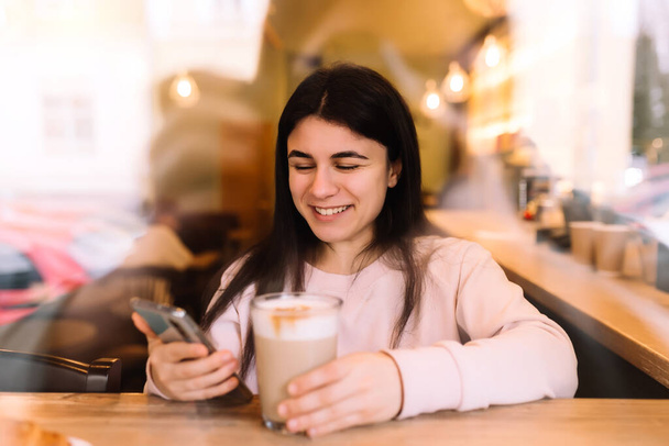 Girl smiling, drinks coffee in cafe and reading phone. Blurred background. High quality photo - Фото, изображение