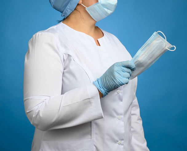 woman doctor in a white medical coat, a disposable mask and a cap stands on a blue background and holds a face mask, close up - Valokuva, kuva