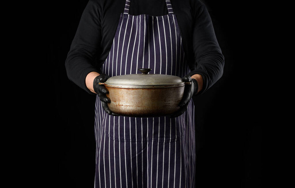 man chef in a striped apron holds an aluminum cauldron with a lid, black background - Fotoğraf, Görsel