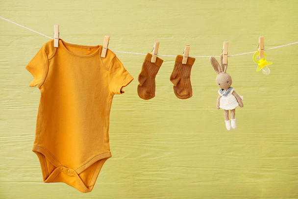 Baby clothes, toy and pacifier hanging on rope against color wooden background - Фото, изображение