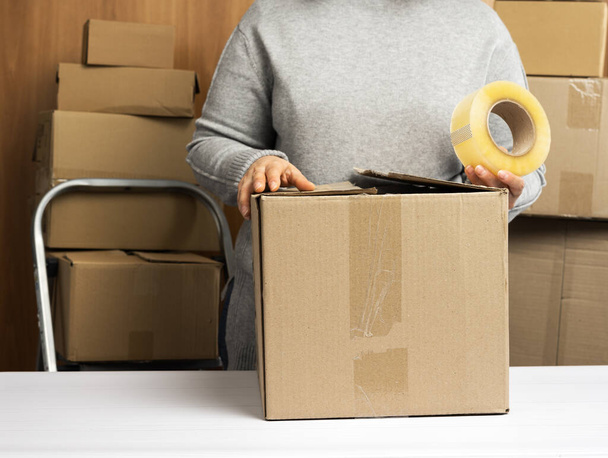 woman in a gray sweater holds a roll of duct tape and packs brown cardboard boxes on a white table, behind a stack of boxes. Moving concept - Foto, afbeelding