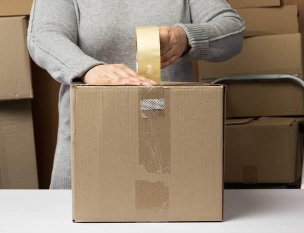 woman in a gray sweater holds a roll of duct tape and packs brown cardboard boxes on a white table, behind a stack of boxes. Moving concept - Foto, Imagem