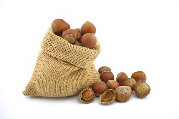 Small hessian bag filled with whole ripe hazelnuts spilling onto a white background - Foto, Imagem