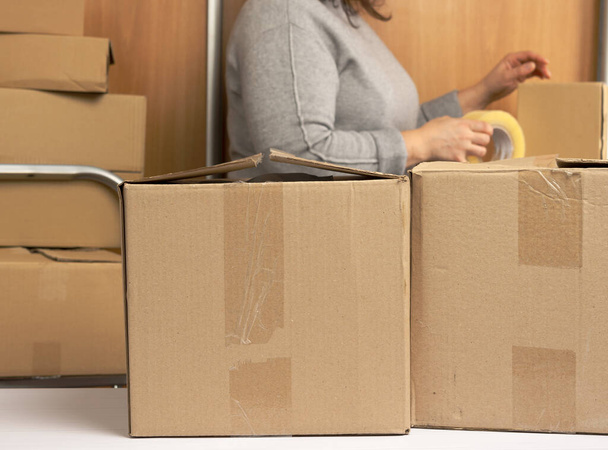 woman in a gray sweater holds a roll of duct tape and packs brown cardboard boxes, behind a stack of boxes. Moving concept - Foto, Bild