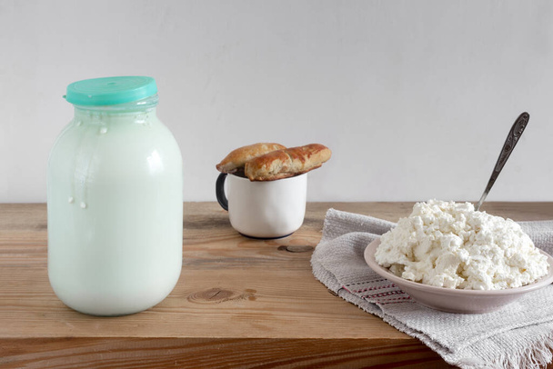 On the wooden table is a glass jar of milk, a plate of homemade cottage cheese and pies. Front view, copy space. - Foto, afbeelding