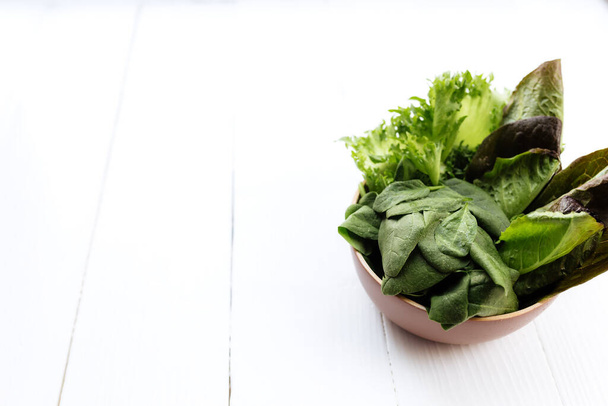 Flat lay with bowl of fresh green salad leaves of spinach and lettuce, romaine and basil on white wooden background. Healthy vegetarian eating concept. High quality photo - Photo, image