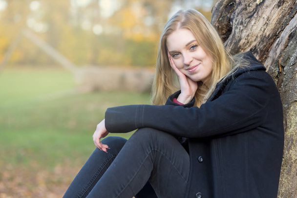 Portrait of smiling girl sitting in autumn park. The girl is looking at the camera. - Valokuva, kuva