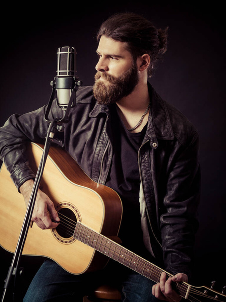 Young bearded man sitting and playing an acoustic guitar over black background.. - Fotoğraf, Görsel