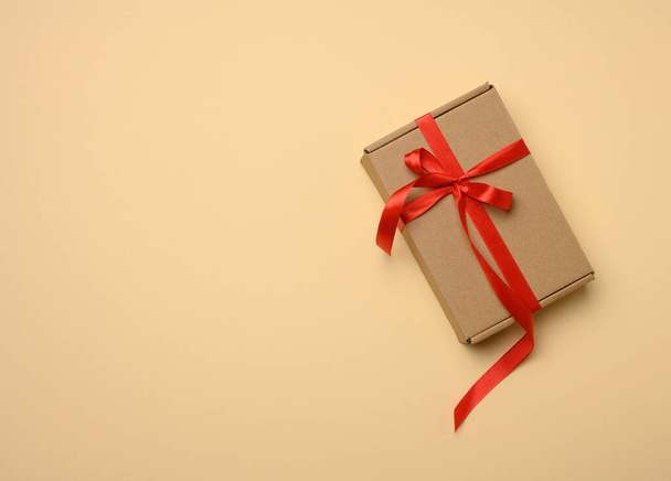 rectangular brown cardboard box tied with a silk red ribbon on a beige background, top view, close up - Фото, изображение