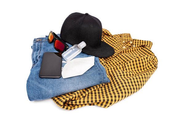 Modern stylish male outfit for the pandemic with jeans, plaid shirt, snapback cap, sunglasses, mobile phone, hand sanitizer and face mask, isolated on white background. The COVID-19 new normal. - Foto, Imagem
