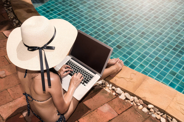 Asian Young Woman using her laptop with big hat on poolside edge on sunny day blue water - Foto, Imagen