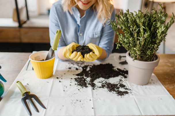 Young woman in gloves holds pile of peat for home plants, florist hobby. Female person takes care of domestic flowers - Foto, afbeelding