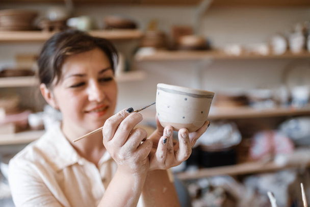 Female master paints a pot, pottery workshop. Woman molding a bowl. Handmade ceramic art, clay tableware - Photo, Image