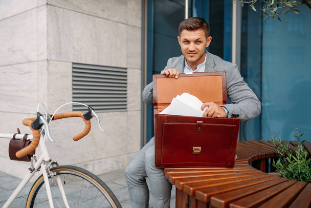 Businessman bicyclist in suit puts document into the briefcase in downtown. Business person riding on eco transport on city street - Fotografie, Obrázek