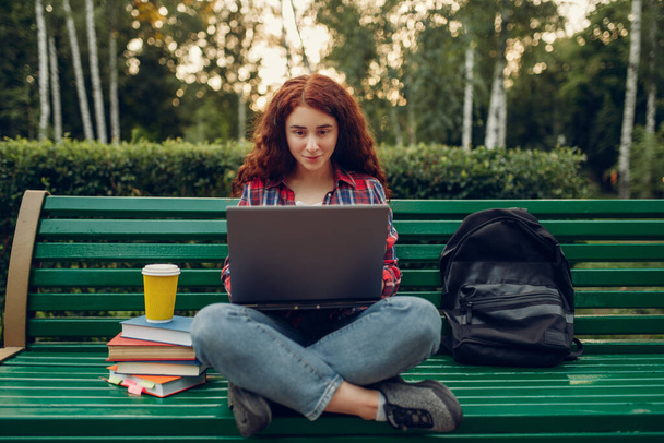 Female student with laptop sitting on the bench in yoga pose, summer park, front view. Ginger teenager leisures outdoors, pretty girl relax outside - Zdjęcie, obraz