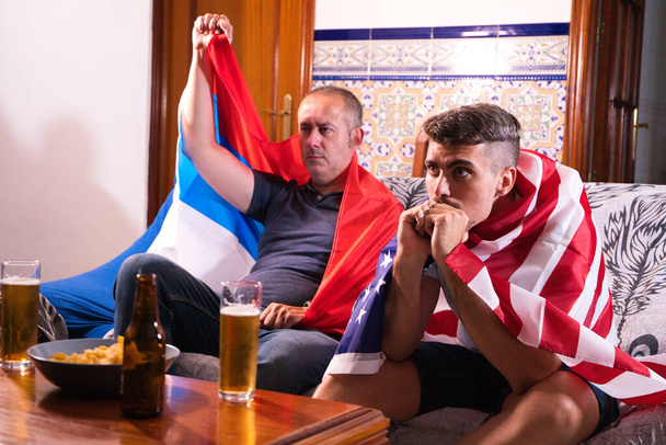 Twee mannelijke vrienden en sportfans kijken naar een sportevenement op televisie terwijl ze een biertje drinken. Elk van de mannen draagt een vlag op zijn schouders. Sportconcept - Foto, afbeelding