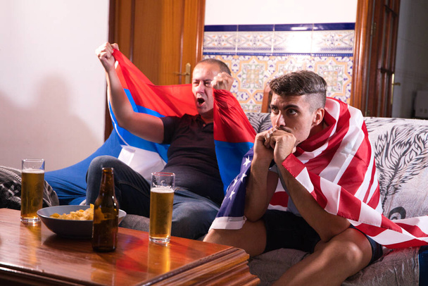 Deux amis masculins et des amateurs de sport regardent un événement sportif à la télévision tout en buvant une bière. Chacun des hommes porte un drapeau sur ses épaules. Concept sportif - Photo, image