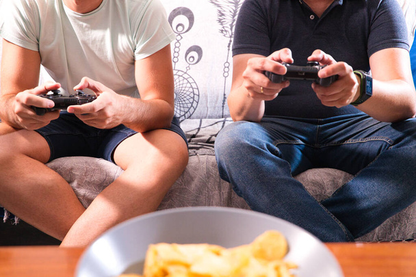 Detalles de las manos de dos amigos sosteniendo el controlador de una videoconsola, los hombres están jugando videojuegos mientras toman una cerveza con bocadillos. Concepto de videojuegos y amistad. - Foto, imagen