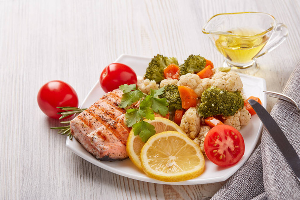 Grilled salmon fillet  garnished with vegetables, lemon, tomato, herbs lying on plate on wooden background. Healthy food. Keto diet. - Foto, Bild