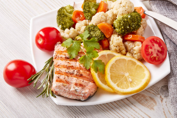 Grilled salmon fillet  garnished with vegetables, lemon, tomato, herbs lying on plate on wooden background. Top view. Healthy food. Keto diet. - Foto, Imagen