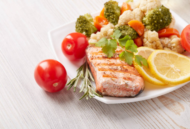 Grilled salmon fillet  garnished with vegetables, lemon, tomato, herbs lying on plate on wooden background. Top view. Healthy food. Keto diet. - Foto, Bild