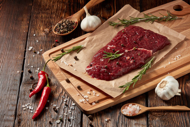 Raw fresh meat. Beef steak on light wooden cutting board, rosemary twigs, spices and vegatables on brown wooden background. Steak menu - Fotó, kép