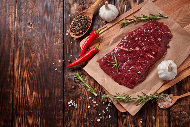 Raw fresh meat. Beef steak on light wooden cutting board, rosemary twigs, spices and vegatables on brown wooden background. Steak menu. Top view with copy space - Photo, Image