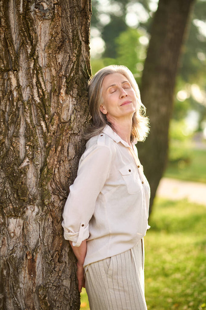 Woman leaning against tree in optimistic mood - Zdjęcie, obraz