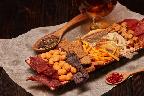 Glass with beer and set of various kind snacks: jerky,  potato chips, salted nuts, stockfish, bread spicy crackers, dried squids  on dark wooden background. Snacks for beer. - Φωτογραφία, εικόνα