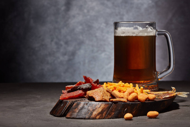 Glass mug with beer and set of various kind snacks: jerky, potato chips, salted nuts, stockfish, bread crackers on brown wooden tray on dark gray textured  background. Snacks for beer. - Photo, image
