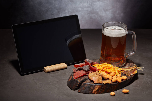 Glass mug with beer, set of various kind snacks and blank computer tablet on dark gray textured  table on grey background. Snacks for beer. - Foto, imagen
