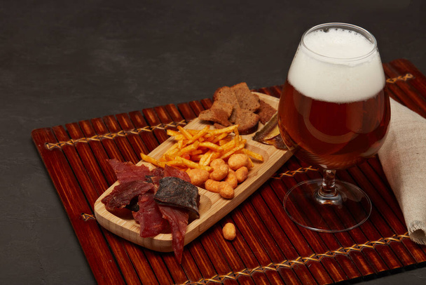 Glass with beer and set of various kind snacks: jerky,  potato chips, salted nuts, stockfish, bread crackers on brown wooden tray on dark gray  background. Snacks for beer. - Zdjęcie, obraz