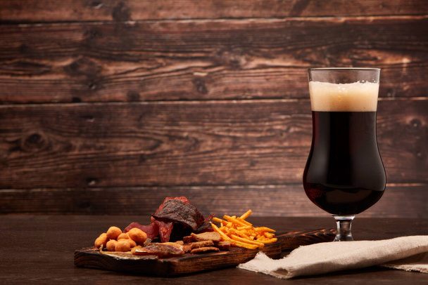 Glass with beer and set of various kind snacks: jerky,  potato chips, salted nuts, stockfish, bread spicy crackers on dark brown wooden background with copy space. Snacks for beer. - Фото, зображення