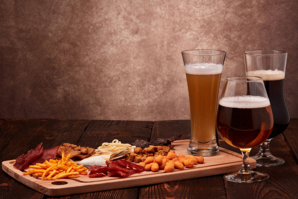 Three glasses with different beer and set of various kind snacks: jerky,  potato chips, salted nuts, stockfish, bread spicy crackers, dried squids on wooden table on brown background. Snacks for beer. - Foto, imagen