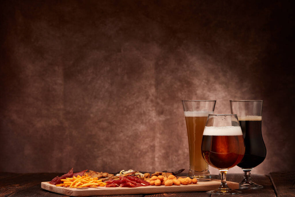 Three glasses with different beer and set of various kind snacks: jerky,  potato chips, salted nuts, stockfish, bread spicy crackers, dried squids on wooden table on brown background. Snacks for beer. - Fotografie, Obrázek