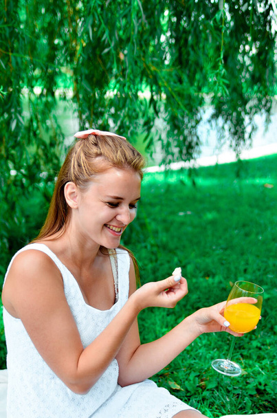 Shot of young girl with hair bow in a white dress and sneakers eating sweets and drink orange juice enjoying outside outdoors park - Foto, Imagen