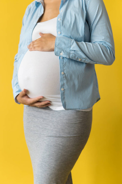Motherhood, femininity, love, care, waiting, hot summer - bright croped Close-up unrecognizable pregnant woman in shirt with small baby shoes hand over tummy rub belly on yellow background, copy space - Foto, Imagem