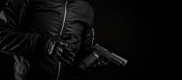 A man in a black jacket and black gloves holding a gun on a dark background. - Foto, Bild