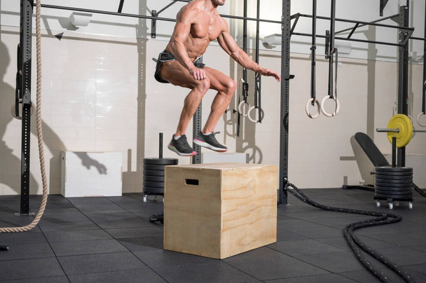 Ο μυώδης αρσενικός αθλητής προπονείται πηδώντας σε ένα ξύλινο κουτί στο σύγχρονο γυμναστήριο. Λειτουργική εκπαίδευση - Φωτογραφία, εικόνα