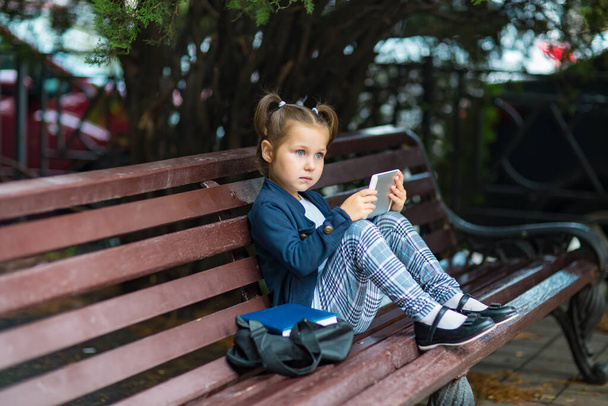 a small, beautiful girl in a school uniform, sitting on a bench, and holding a tablet in her hands - Zdjęcie, obraz